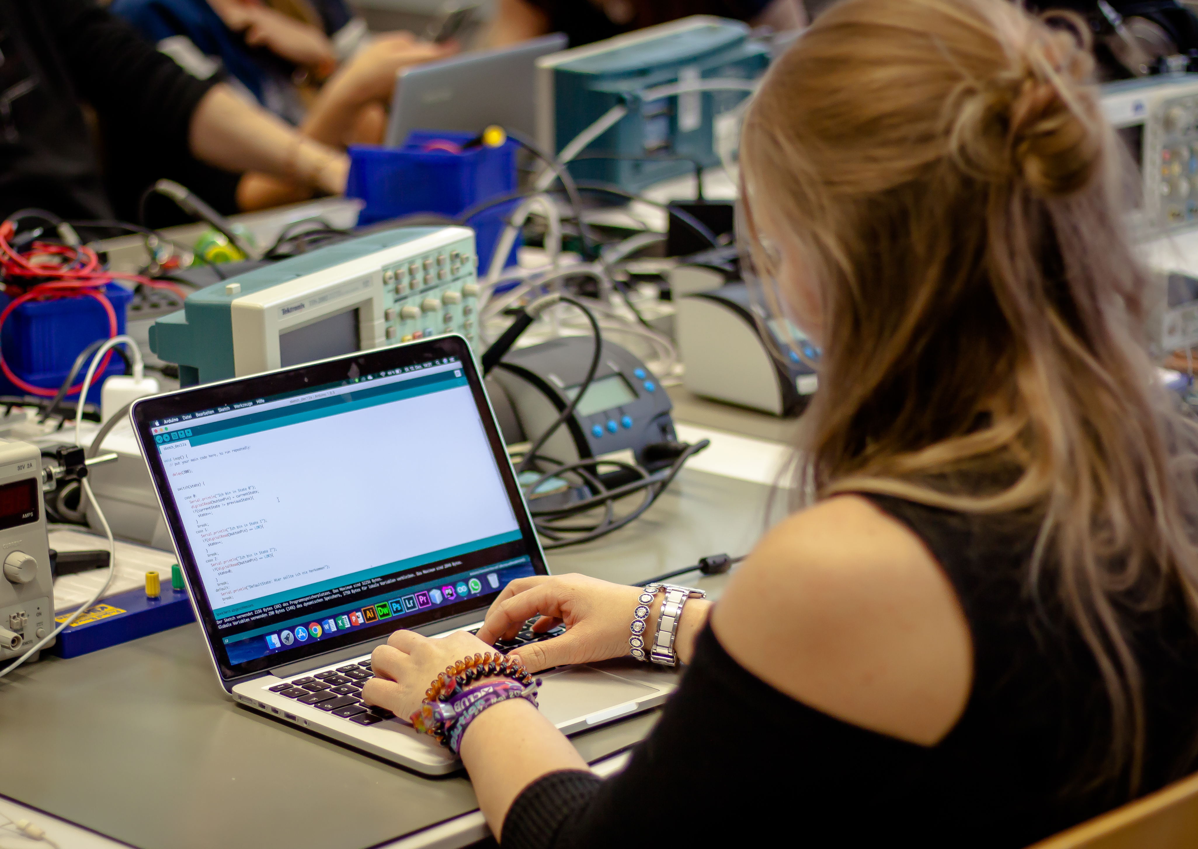 Bild von einer Person, welche an einem Laptop programmiert. Im Hintergrund sind elektronische Geräte, Kabel und eine Lötstation zu sehen